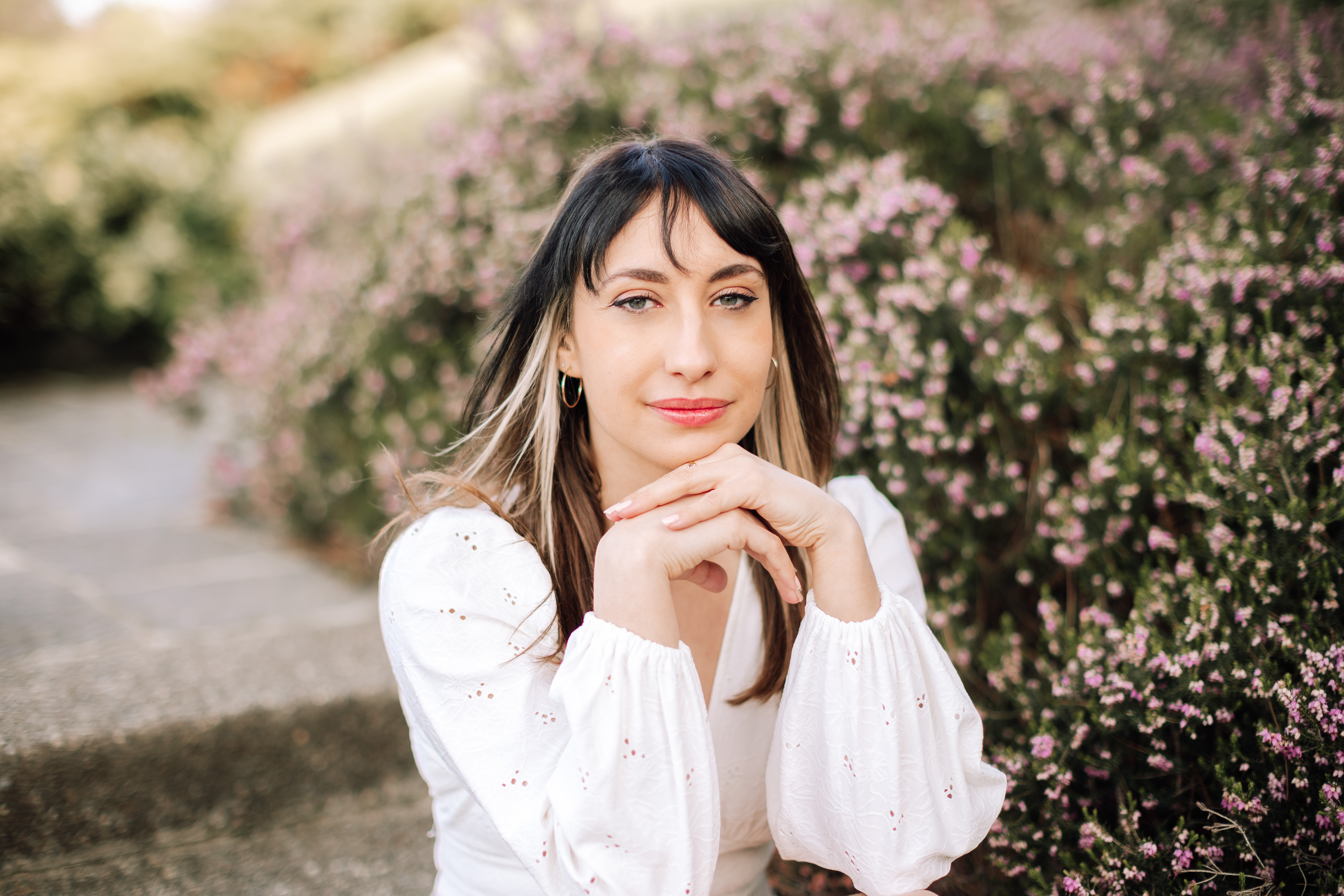 Velia Ferracini, brune aux longs cheveux laissés libres qui descendent jusqu'à sous ses clavicules, sur ses épaules et dans son dos, pose vêtue d'un pull blanc, sourire et regard doux, face à l'objectif. Ses mains sont posées l'une sur l'autre un peu sous son menton, avant bras repliés en direction de son visage. L'arrière-plan est flouté, mais on devine un arbuste en fleur derrière elle à sa droite.
