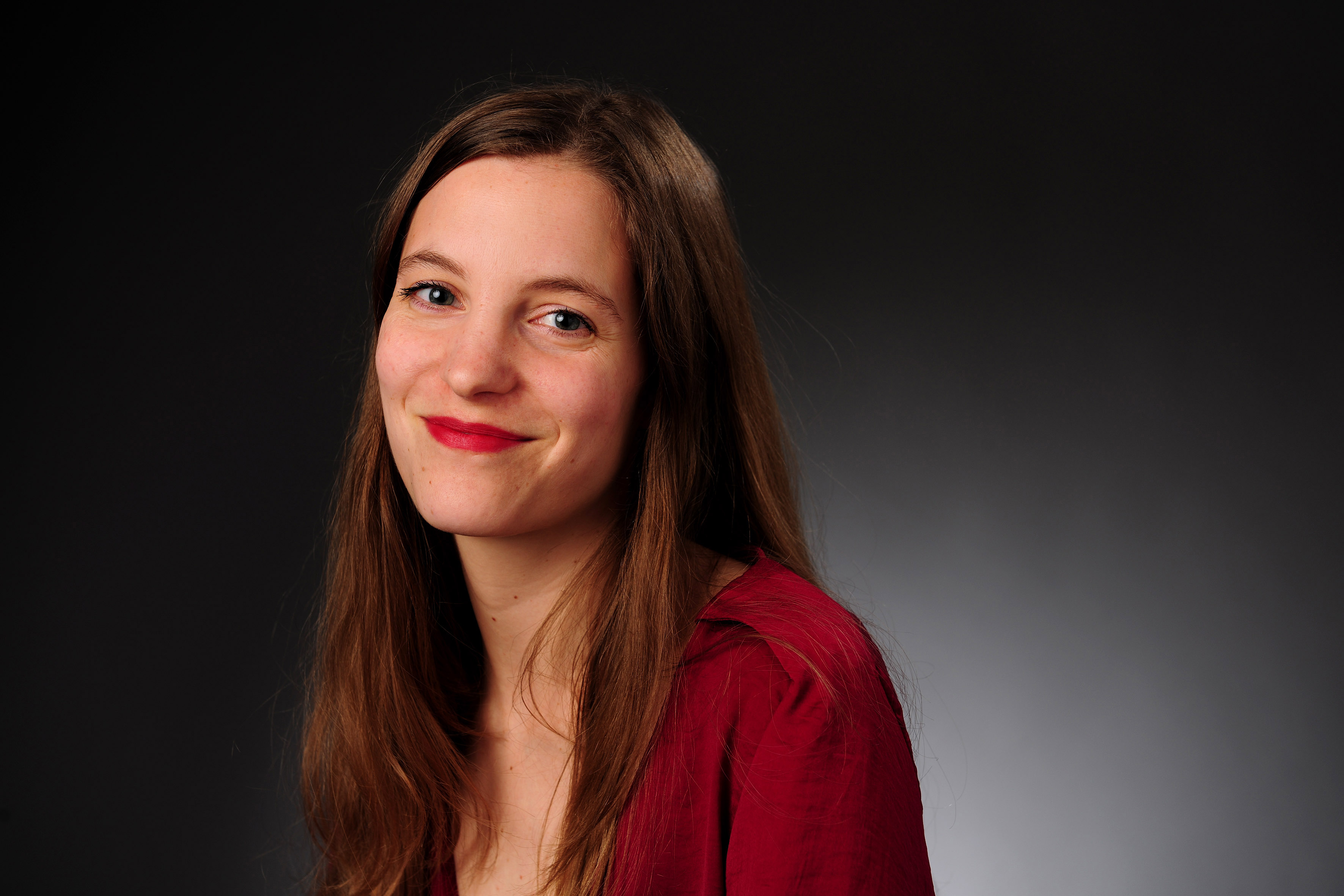 Lolvé Tillmanns brune aux longs cheveux pose en chemisier rouge foncé sur fond noir.