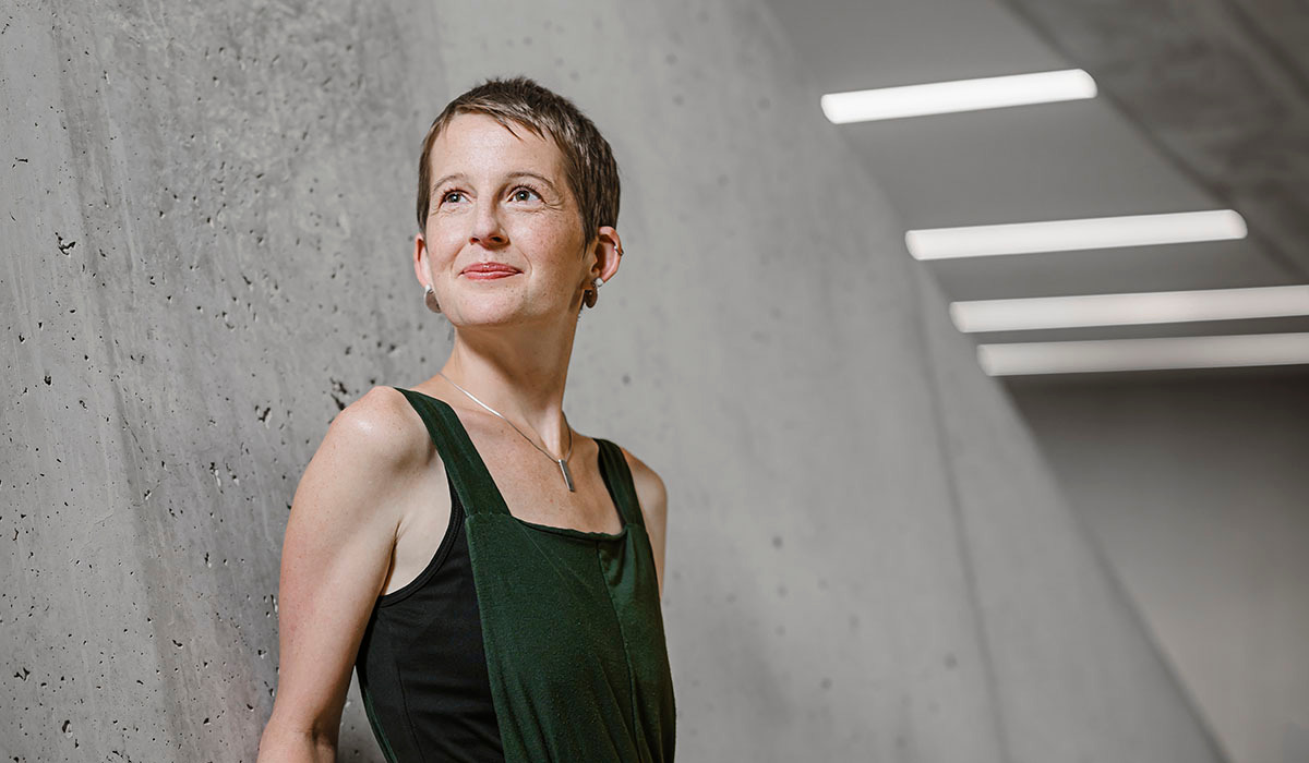 Brune aux cheveux très courts, Chloé Falcy pose debout devant un mur en béton dans un décor épuré. Légèrement de profil, souriante bouche fermée, le regard tourné vers la gauche, nimbé de lumière.